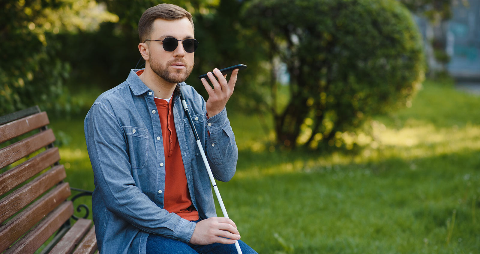 Ein junger Mann mit Langstock und schwarzer Brille spricht in ein Smartphone